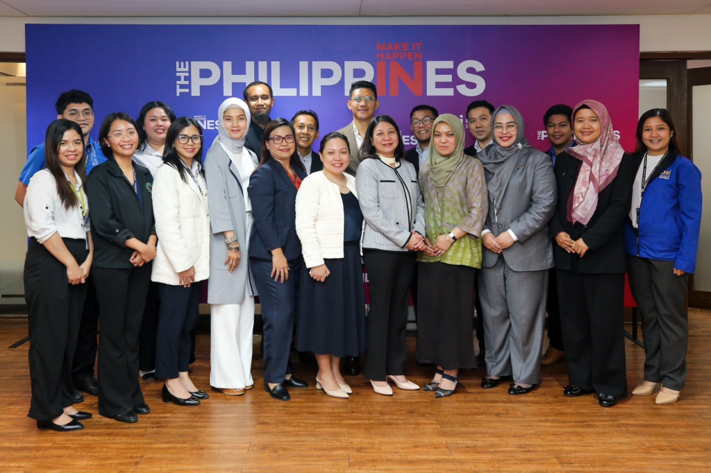 A group of individuals from PTIC in Jakarta, PT Pertamina Gas Negara, and the Philippine Board of Investments stand together in front of a large backdrop that reads “The Philippines” and “Make It Happen.” They wear business attire (some in hijabs) and smile for the camera in a wood-floored room.