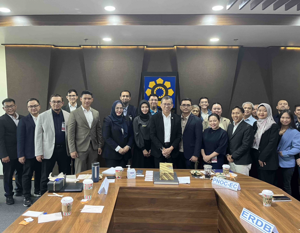 A large group of individuals from PTIC in Jakarta, the Department of Energy, and Pertamina Gas Negara stand together in a conference room. They are dressed in business attire, smiling at the camera, with a DOE emblem displayed on the wall behind them. Tables in front hold nameplates, cups, and documents.