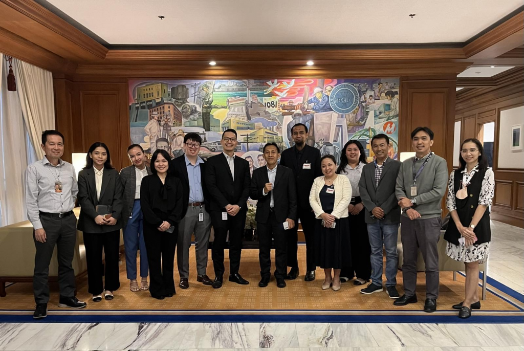 A group of individuals from PTIC in Jakarta, PT Pertamina Gas Negara, and Meralco stand in a well-lit room in front of a large, colorful mural. They are dressed in business attire, smiling, and appear to be gathered for a meeting in November 2024.