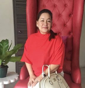 Madonna Elizaga, a woman entrepreneur, sits on a red high-back tufted chair, smiling warmly at the camera. She is wearing a red blouse and holding a beige handbag. A potted plant and a small side table are visible in the background, adding a cozy and elegant touch to the setting.