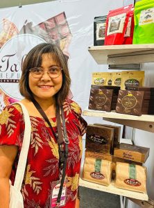 A smiling woman in a red floral blouse stands beside a display shelf featuring “ATBANG” chocolate products, with a booth banner visible in the background.