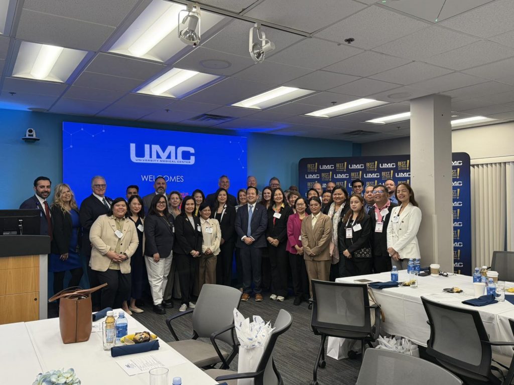 Group photo of diverse professionals in business attire at the UMC University Medical Center. A screen displays the UMC logo and "Welcomes" message. Tables with water bottles are in the foreground.