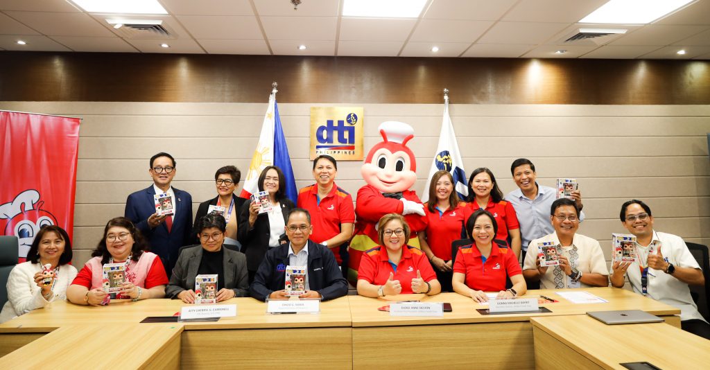 A group of Department of Trade and Industry (DTI) and Jollibee Foods Corporation representatives are in a conference room, holding Jollibee Funko Pop figures. They are posing for a photo during a partnership event with the Philippine flag and the DTI logo in the background, and Jollibee mascot standing with them.