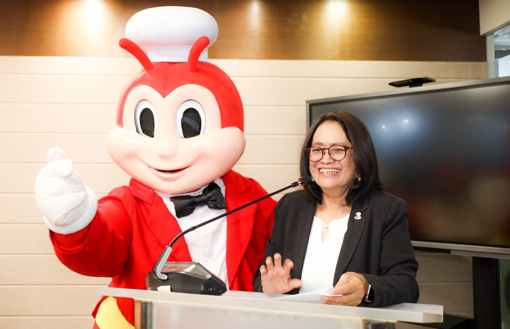 DTI Undersecretary for Regional Operations Group Blesila Lantayona smiles as she speaks at a podium during an event. Standing next to her is the Jollibee mascot, giving a finger heart. A large monitor is behind them.