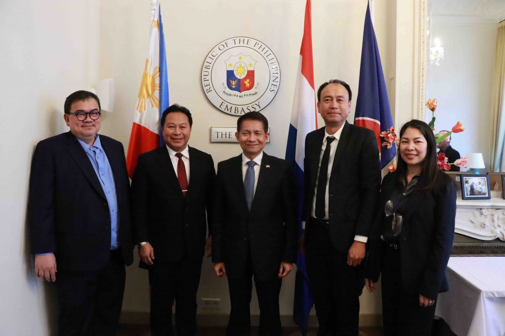 A group photo of five individuals standing in front of the Philippine Embassy seal in the Netherlands, with the Philippine and Dutch flags displayed behind them. Senior Undersecretary Emil K. Sadain of the DPWH (second from left), Philippine Ambassador to the Netherlands Eduardo J. Malaya (center), and Commercial Counsellor Magnolia Misolas-Ashley, PTIC-Brussels (rightmost) are among the delegates. The setting includes a formal indoor space with floral decorations and framed photographs.