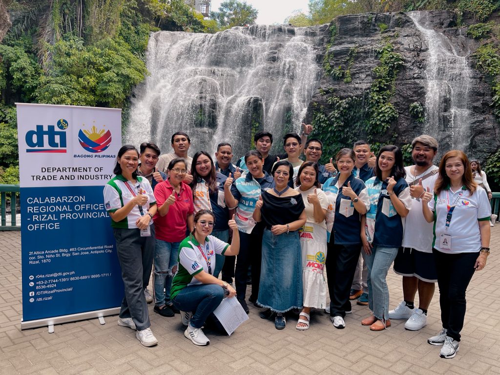 A group photo of DTI CALABARZON - Rizal Provincial Office staff in front of a waterfall. They are holding a banner with the DTI logo and office information and giving thumbs-up gestures. This suggests a team outing or activity at a natural landmark.