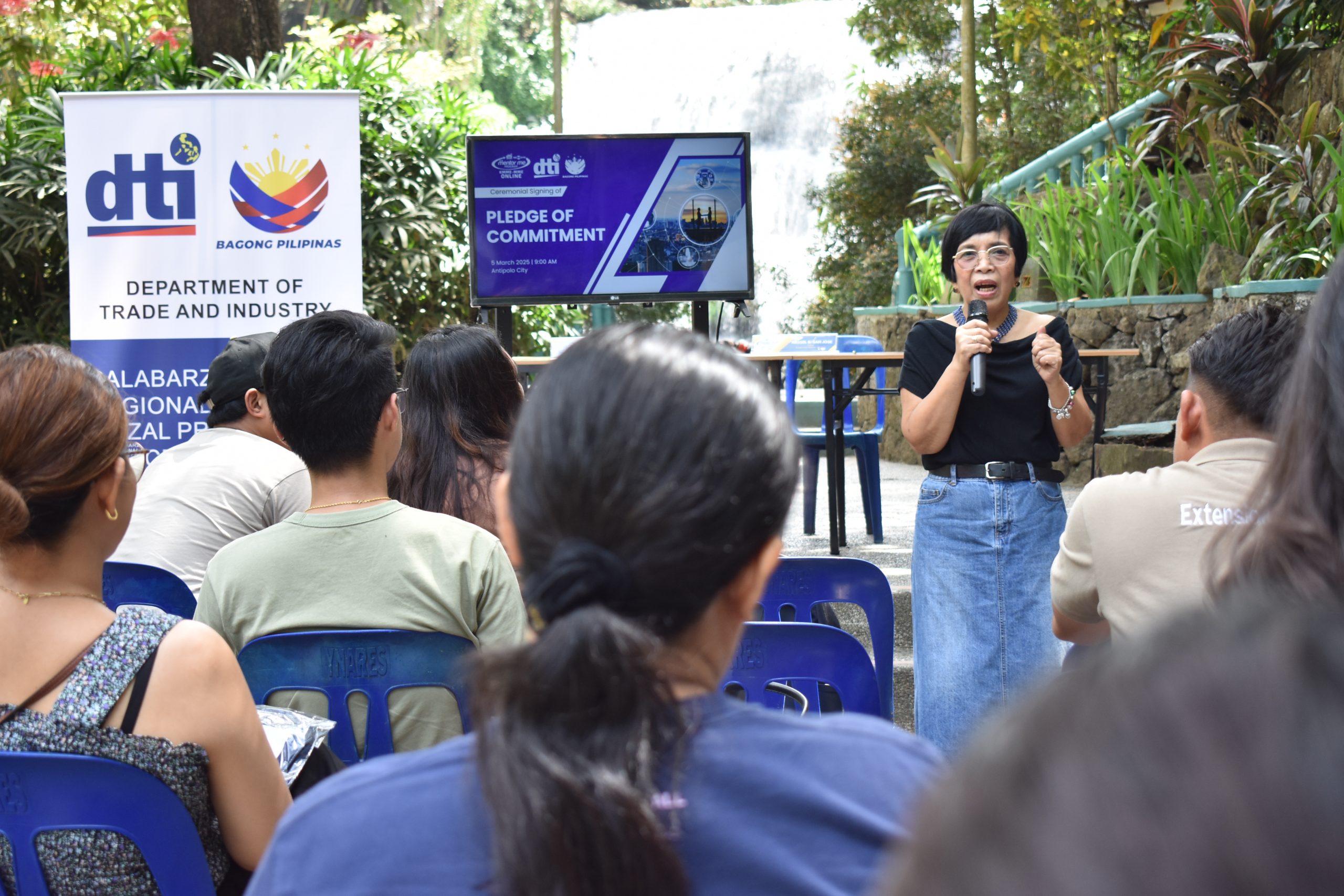 A DTI event taking place outdoors, with a woman addressing a group. The event includes a "PLEDGE OF COMMITMENT" presentation, as shown on the screen, and the DTI CALABARZON banner is visible. The location features a waterfall, suggesting an outdoor or nature-related gathering.