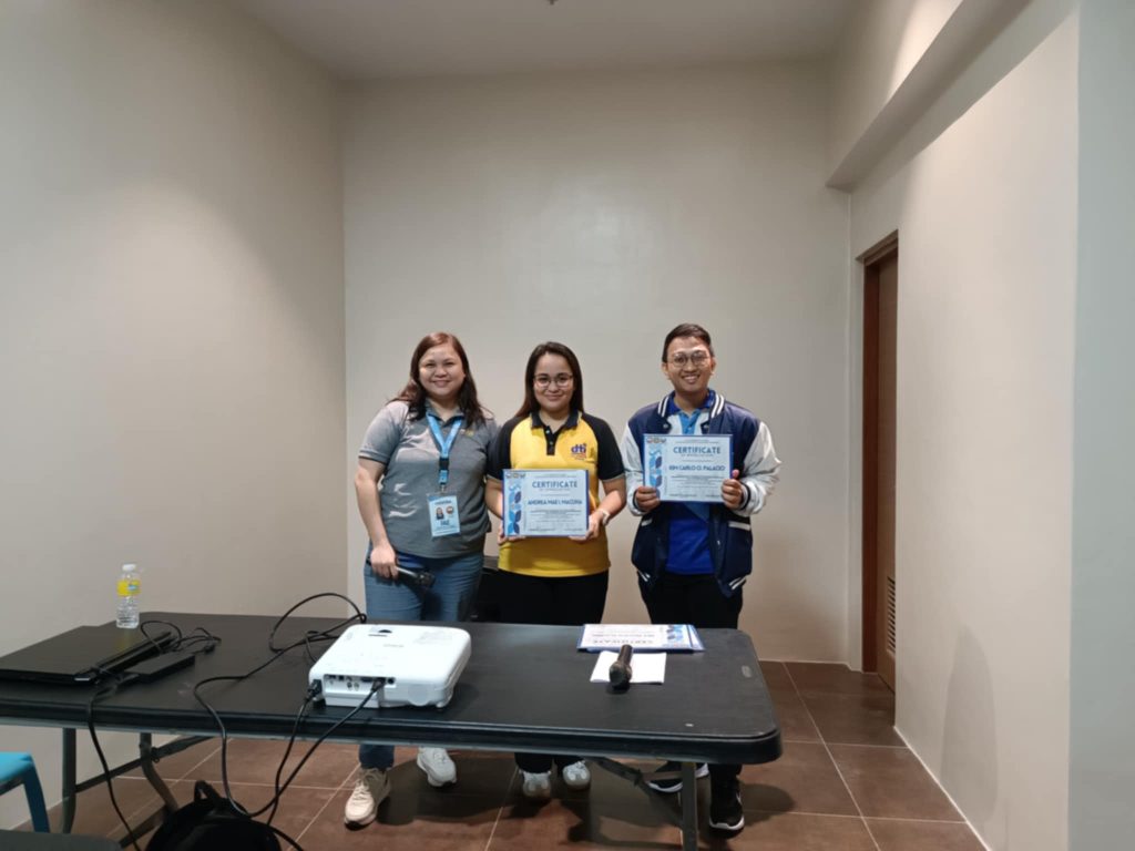Three individuals are standing in a room, each holding a certificate. A table with a laptop, projector, and other equipment is in the foreground. The walls are plain and painted a light color.