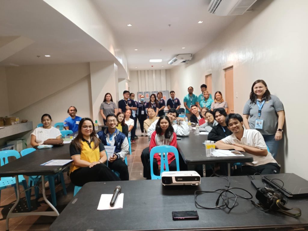 A group of people are gathered in a room with tables and chairs. A projector is set up at the front, suggesting a presentation or training session. Some individuals are looking at the camera, while others are looking forward.