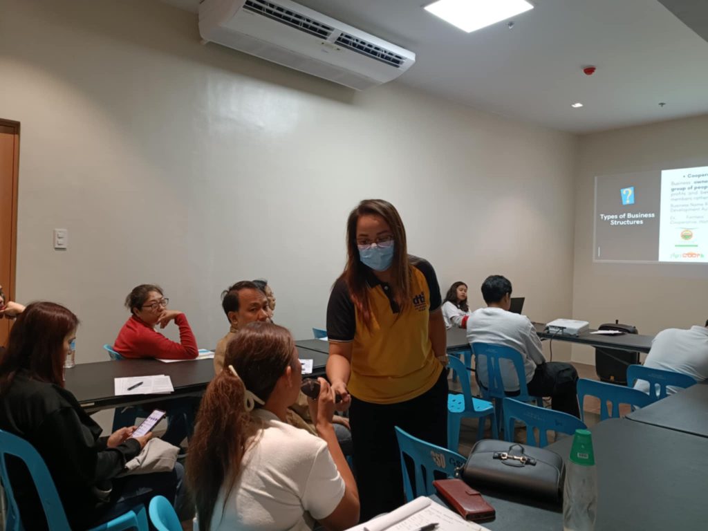 Attendees participate in a business training or seminar, as indicated by the presentation on "Types of Business Structures." A facilitator wearing a face mask is interacting with participants. The room appears to be a training or conference space.