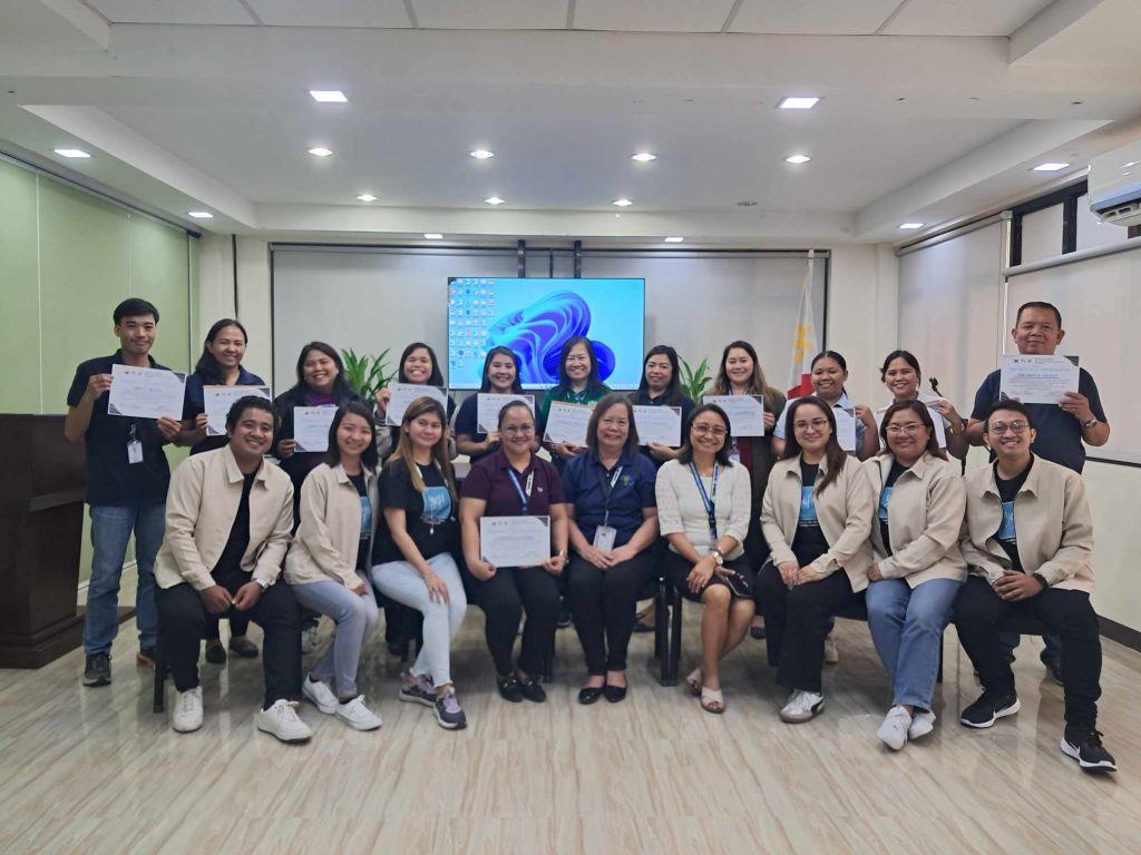A group of 18 people are posing for a photo indoors, holding certificates. They are diverse in age and appear to be celebrating an achievement or graduation. A large screen displaying a Windows logo is visible in the background. The room has a modern interior with light-colored flooring and paneling. The people are dressed in a mix of casual and semi-formal attire.