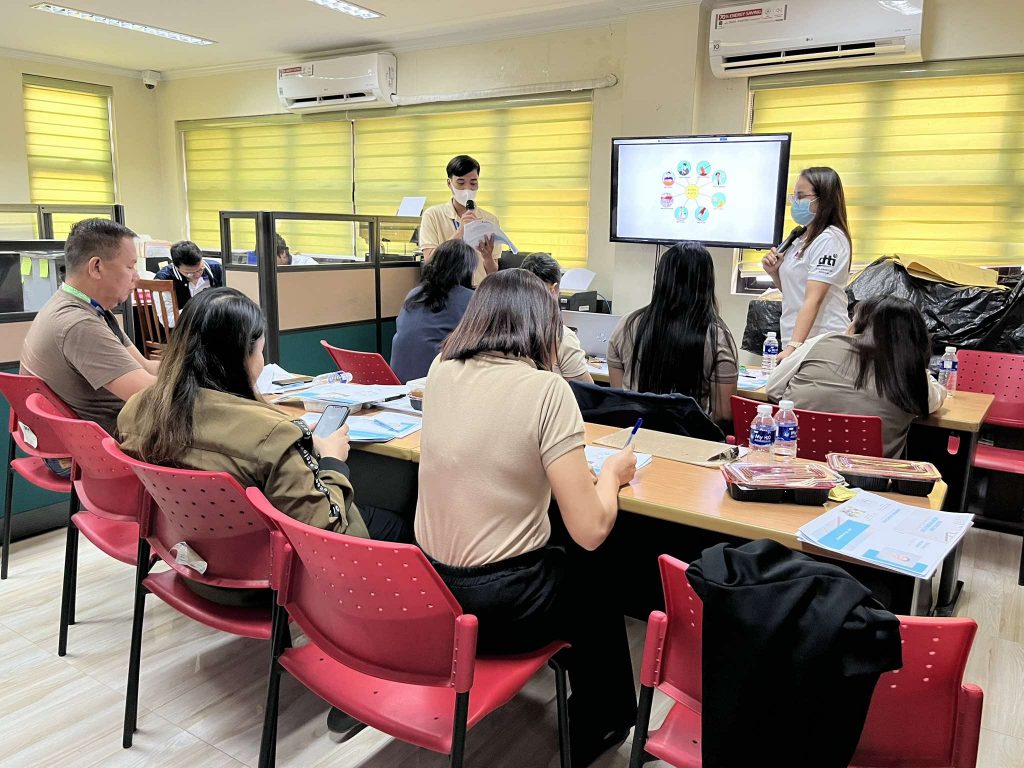 A group of people are seated at tables in a room, facing a screen displaying a graphic. Two individuals appear to be presenting or leading the session, one speaking into a microphone. The room has yellow blinds, cubicles, and air conditioning units on the walls. The participants are seated in red plastic chairs and appear to be taking notes or using their phones. The screen displays a graphic with circular icons. There are water bottles and containers on the tables.