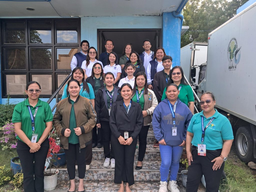 A group of approximately 18 people are posing for a photo on a set of outdoor steps leading up to a building with blue and white accents. The people are diverse in age and appearance, and many are wearing lanyards and green or blue polo shirts. A white truck with a logo is visible in the background. Plants are visible on the steps. The weather appears to be sunny.