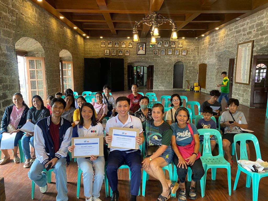 Participants of the DTI-Laguna Financial Literacy and Consumer Education seminar gather for a group photo inside the venue in Pagsanjan, Laguna. Three participants seated in the front row proudly hold up their certificates of completion/participation.
