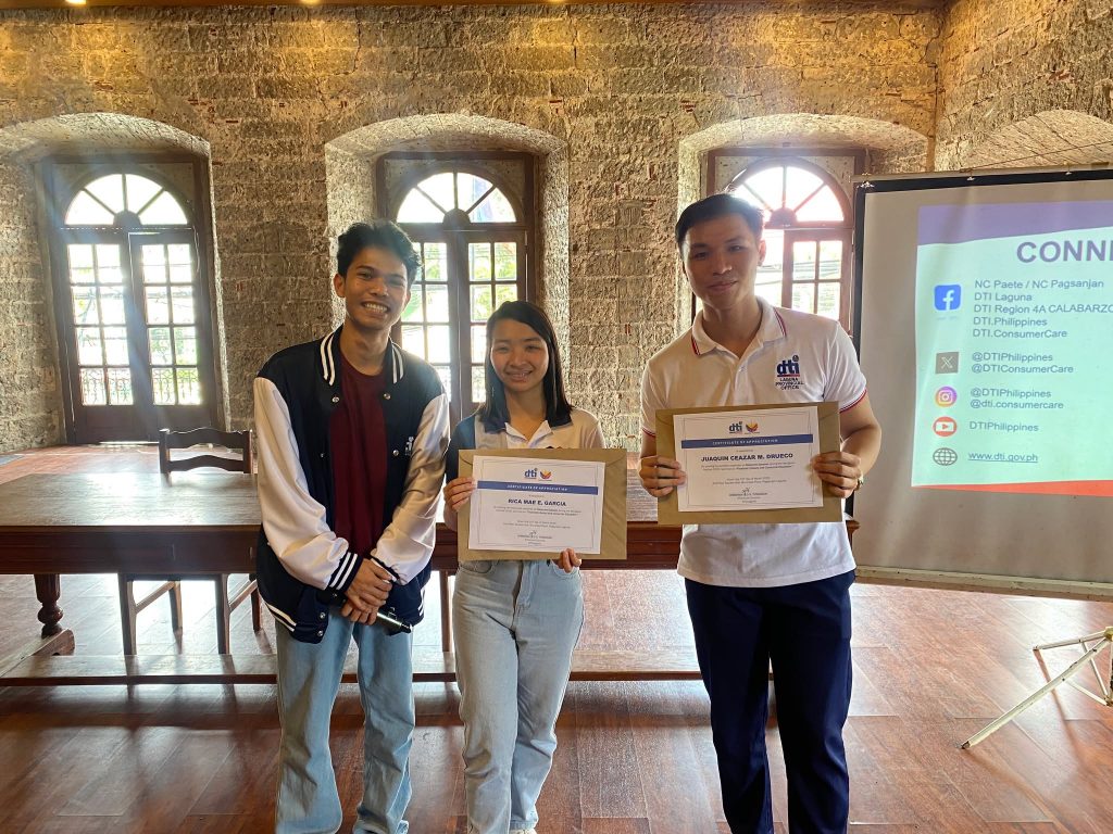 Three participants stand together holding their certificates after completing the DTI-Laguna seminar on Financial Literacy and Consumer Education in Pagsanjan, Laguna. They are posing inside the event venue, with a presentation screen visible in the background.