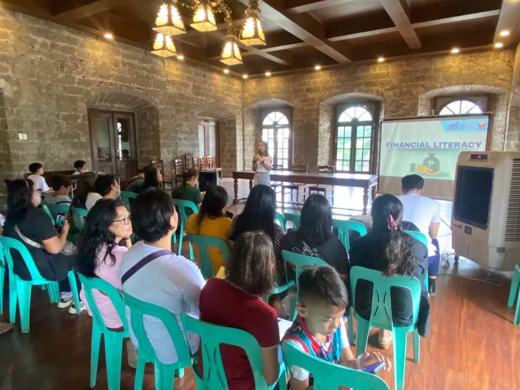 A speaker delivers a presentation on "Financial Literacy," visible on the projector screen, to participants seated during the DTI-Laguna seminar for DSWD SLP beneficiaries in Pagsanjan, Laguna.