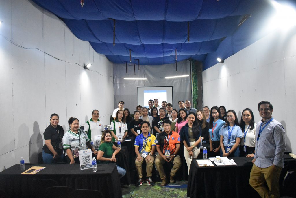 A wide shot of a group of approximately thirty individuals, appearing to be a mix of men and women, posing for a photo at a workshop. They are standing and sitting behind tables in a room with white walls and a blue draped ceiling. A screen displaying a presentation is visible in the background.