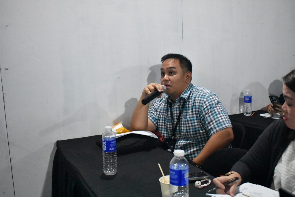 A man is speaking into a microphone while seated at a table. There are water bottles on the table.
