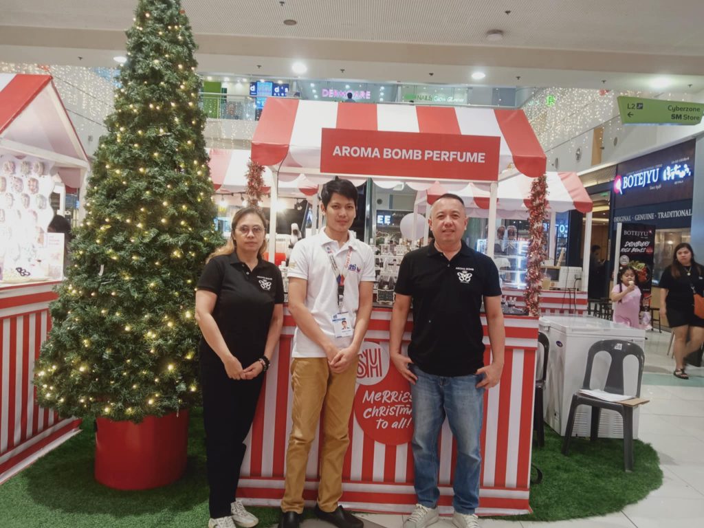 Three individuals, two men and one woman, stand in front of a perfume kiosk labeled "AROMA BOMB PERFUME" within a shopping mall setting. A Christmas tree is positioned to the left of the kiosk.