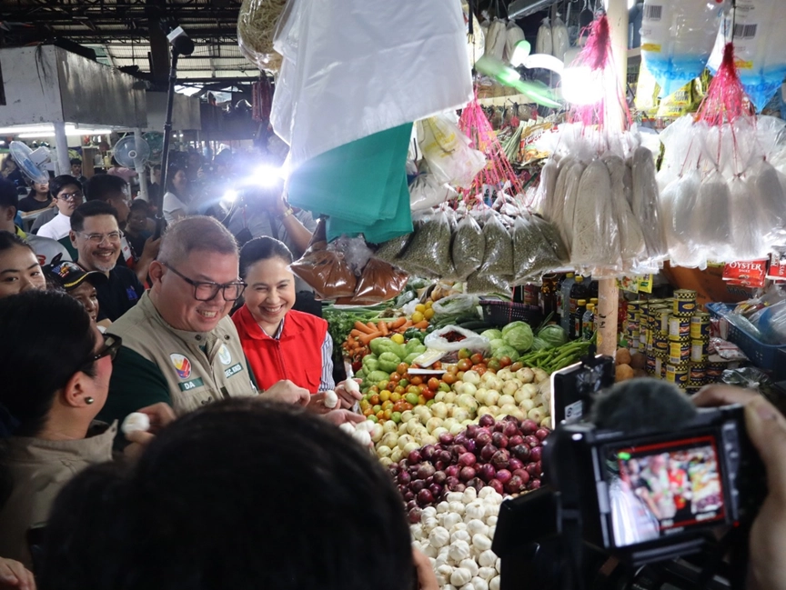 DTI and DA officials, DTI Secretary Cristina A. Roque and DA Secretary Francisco Tiu Laurel Jr., inspecting produce at a Quezon City market during a price and supply monitoring operation. The scene shows vegetables and the presence of media documenting the event.