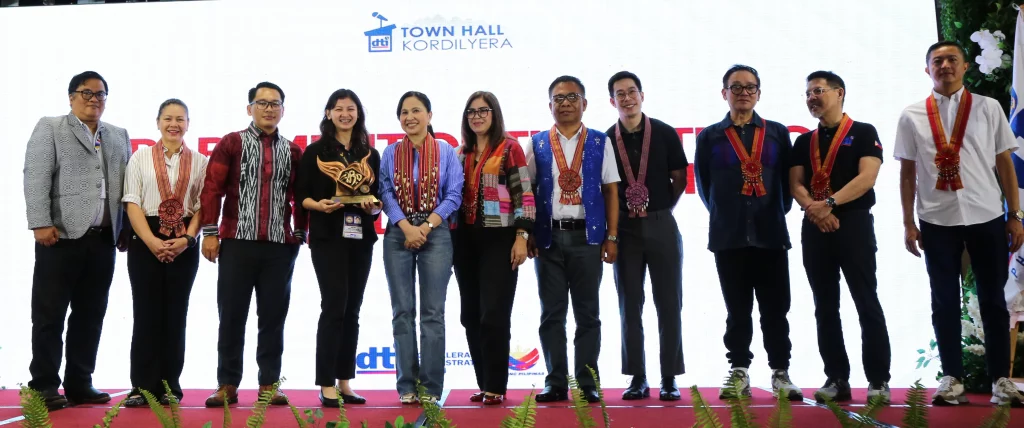 A group of DTI officials and awardees pose for a photo on stage during the DTI Town Hall Kordilyera recognition ceremony in Baguio City. Several individuals wear medals or sashes, some with Cordilleran patterns, and one woman holds a small trophy or plaque. The DTI official presumed to be Secretary Roque stands fifth from the left, wearing a woven vest/sash.