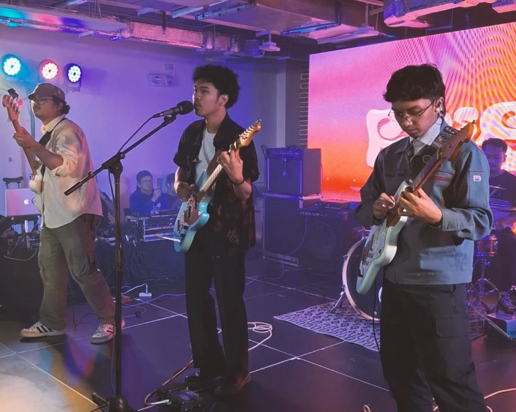 A band with a bassist, singer/guitarist, and guitarist performs live on stage under blue and purple lights during the culminating concert for the DTI-MINT College Malikhaing Pinoy: Music Business Masterclass.