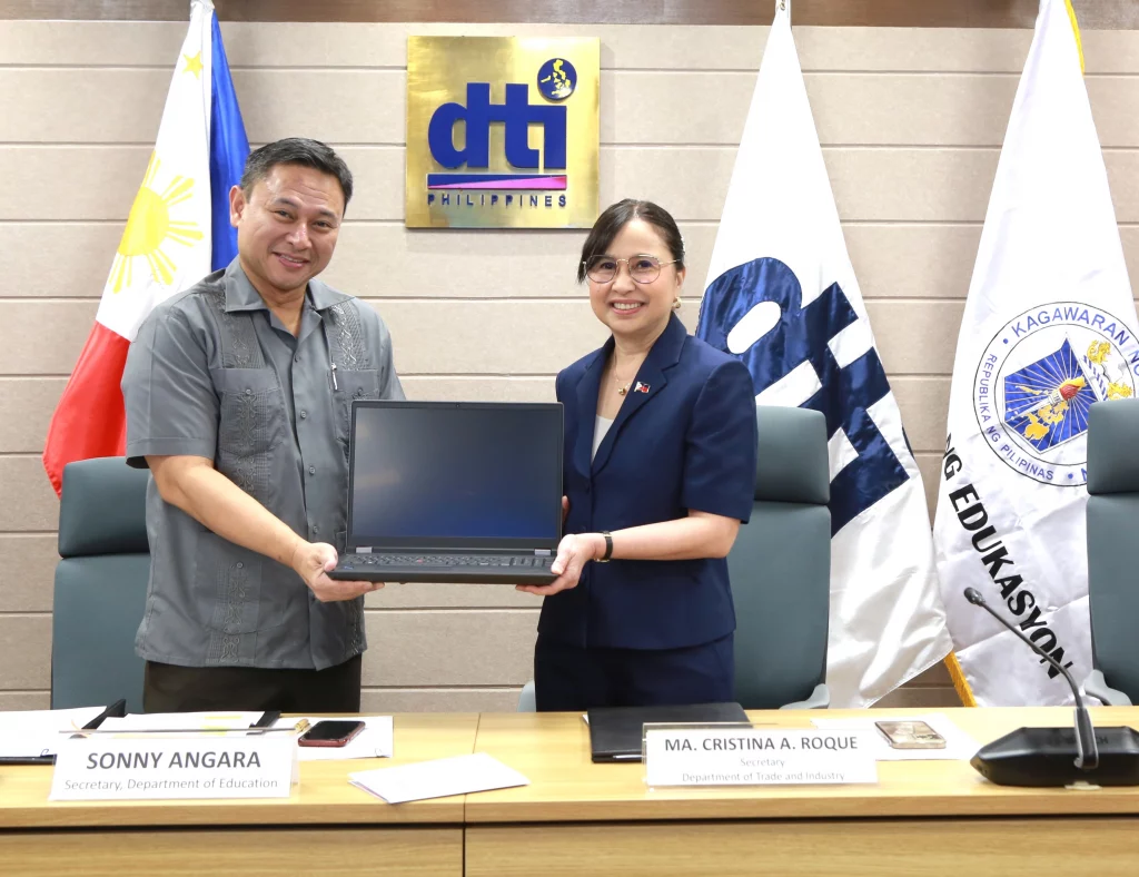DTI Secretary Cristina A. Roque hands over a laptop to DepEd Secretary Sonny Angara, symbolizing a partnership between the Department of Trade and Industry (DTI) and the Department of Education (DepEd) to integrate artificial intelligence (AI) into the Philippine education system. The DTI and DepEd logos are visible in the background, along with the Philippine flag.