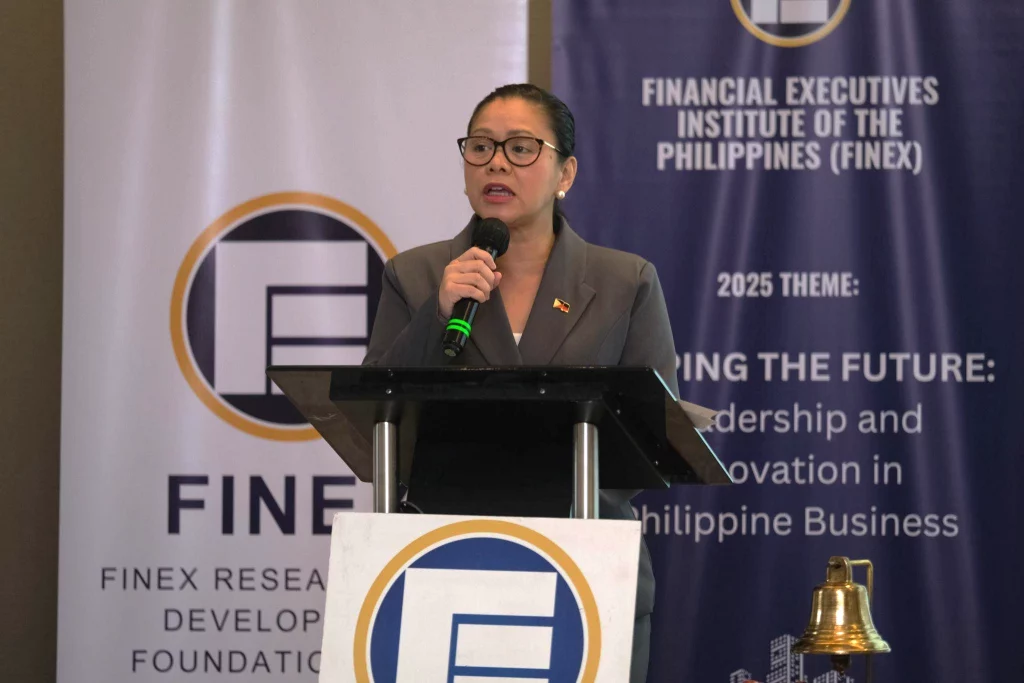 Secretary Cristina Aldeguer-Roque speaks into a microphone at a podium during a Financial Executives Institute of the Philippines (FINEX) event, with banners displaying the organization's name and the 2025 theme visible behind her.