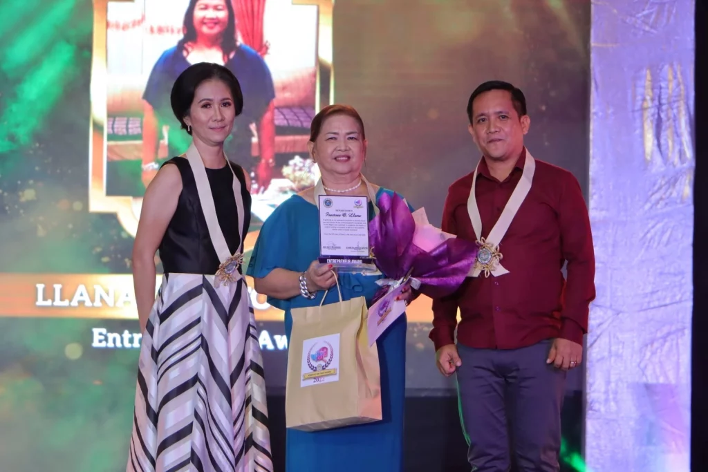 Three individuals are on a stage. A woman in the center, Fructosa Llana, is holding an award and a bouquet of flowers, and has a shopping bag over her shoulder. Two other individuals, a woman on her left and a man on her right, are standing beside her, also wearing award ribbons around their necks. A screen in the background displays text and an image.