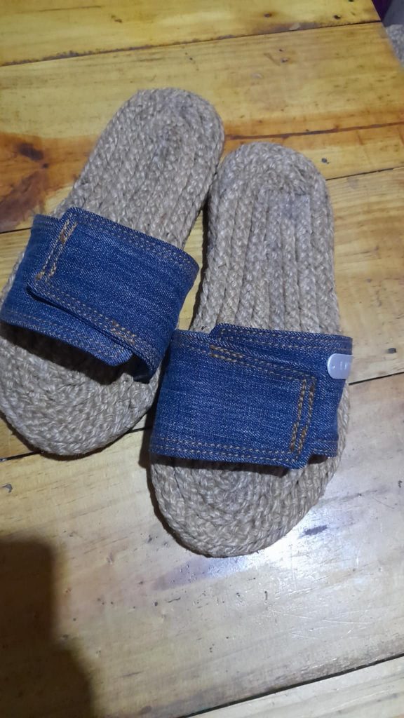 A pair of handmade slippers is shown on a wooden surface. The slippers have thick soles made from tightly coiled jute or a similar natural fiber. The straps are made of blue denim fabric with visible stitching. The slippers have a simple, rustic design. Given the likely origin based on previous images and the current location, these could be Filipino-made.