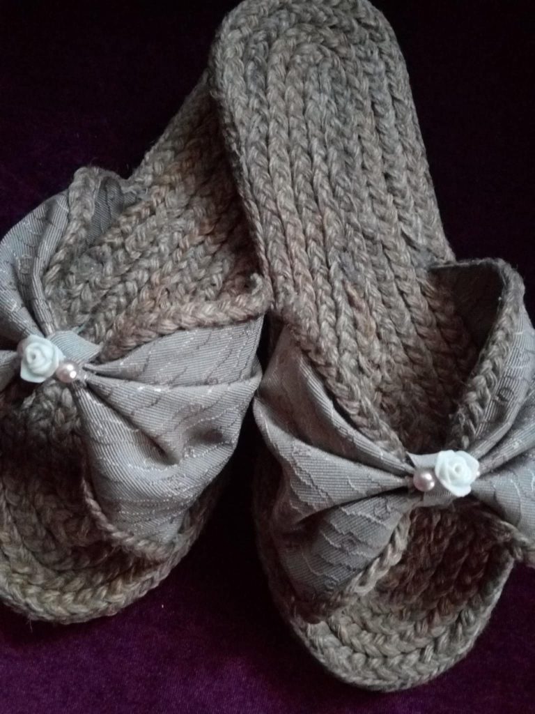 A close-up shot features a pair of handmade slippers resting on a dark purple surface. The slippers have thick, textured soles made from tightly braided jute or a similar natural, coarse fiber. The upper part of each slipper is adorned with a wide, light gray fabric bow that has a subtle leaf-like pattern. At the center of each bow is a small, off-white decorative element resembling a flower or bead. The slippers have a rustic yet elegant appearance, suggesting they are handcrafted. Given the likely origin based on previous images, these could be Filipino-made.