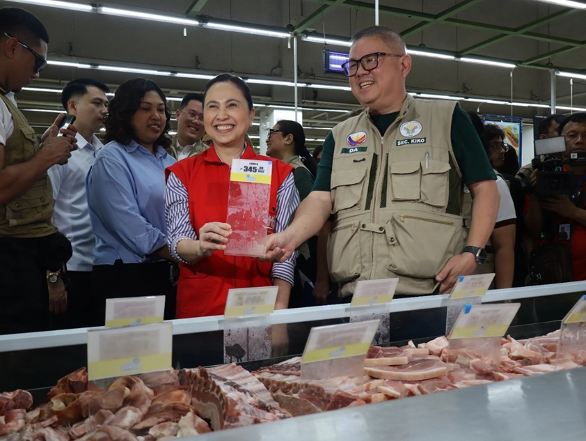DTI and DA officials including Secretary Roque inspecting meat products at a market or supermarket during a price monitoring activity. The woman is holding up a price tag, illustrating the focus on pricing. The scene suggests an effort to ensure compliance with SRP/MSRP.