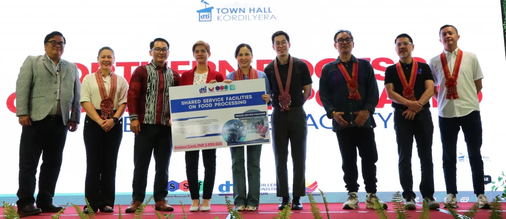 DTI Secretary Cristina A. Roque (center) holds a ceremonial presentation board detailing "Shared Service Facilities on Food Processing", flanked by officials and industry partners wearing medals/sashes on stage during the DTI Town Hall Kordilyera recognition event in Baguio City.