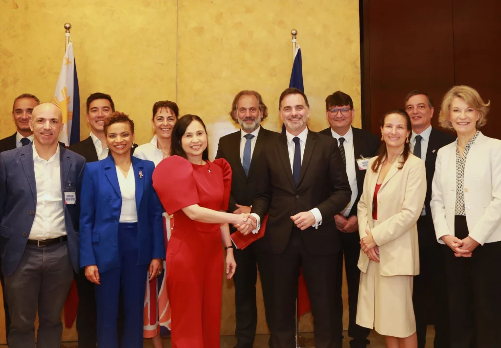 DTI Secretary Cristina A. Roque, in a red Filipiniana pantsuit, shakes hands with a male French delegate (likely Minister Laurent Saint-Martin) during the Philippines-France Business Forum, posing for a group photo with other Filipino and French officials in front of the Philippine and French flags.