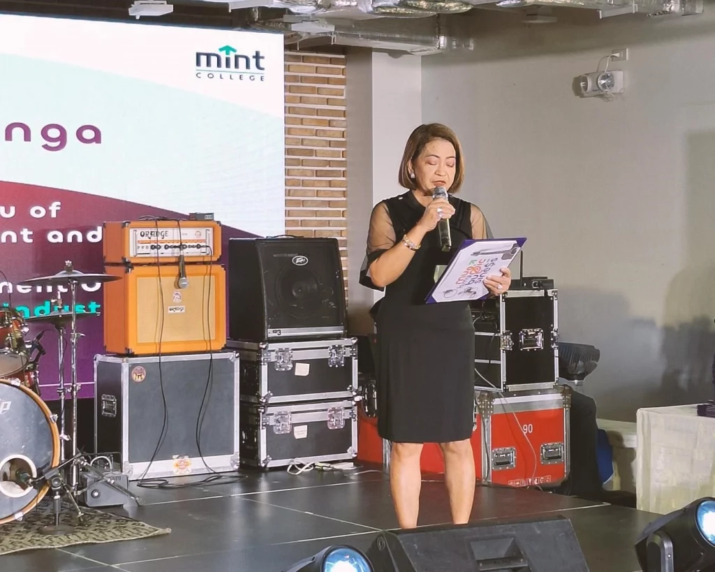 DTI Creative Industries Development Office OIC Director Lilian Salonga, wearing a black dress, delivers her speech on stage holding a clipboard and speaking into a microphone during the Malikhaing Pinoy: Music Business Masterclasses culminating event at MINT College.