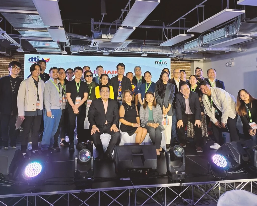 Industry stakeholders, mentors, DTI, and MINT College representatives pose together for a group photo on stage, with DTI and MINT logos visible on the screen behind them, during the Malikhaing Pinoy: Music Business Masterclass culminating concert and networking event.