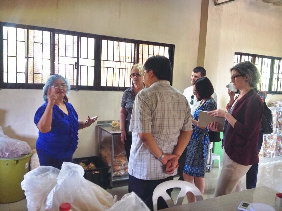 A group of people, likely including Fructosa Llana (the woman in the blue top and hairnet), are gathered inside what appears to be the Frux Food Products facility in Panabo City, Davao del Norte, Philippines. They seem to be in discussion, possibly during a visit or tour related to Fructosa's entrepreneurial success.
