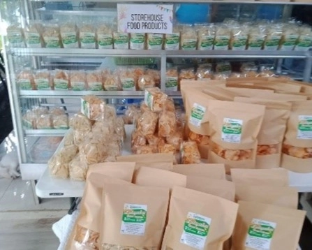 A display shelf showcases various packaged food products from Storehouse Food Products. The sign at the top reads "STOREHOUSE FOOD PRODUCTS." Different types of chips and snacks in clear and brown packaging are arranged on the shelves. In front of the shelf, more packaged products, including what appear to be the LUTYAKO Taro Chips in brown bags, are displayed on a table.