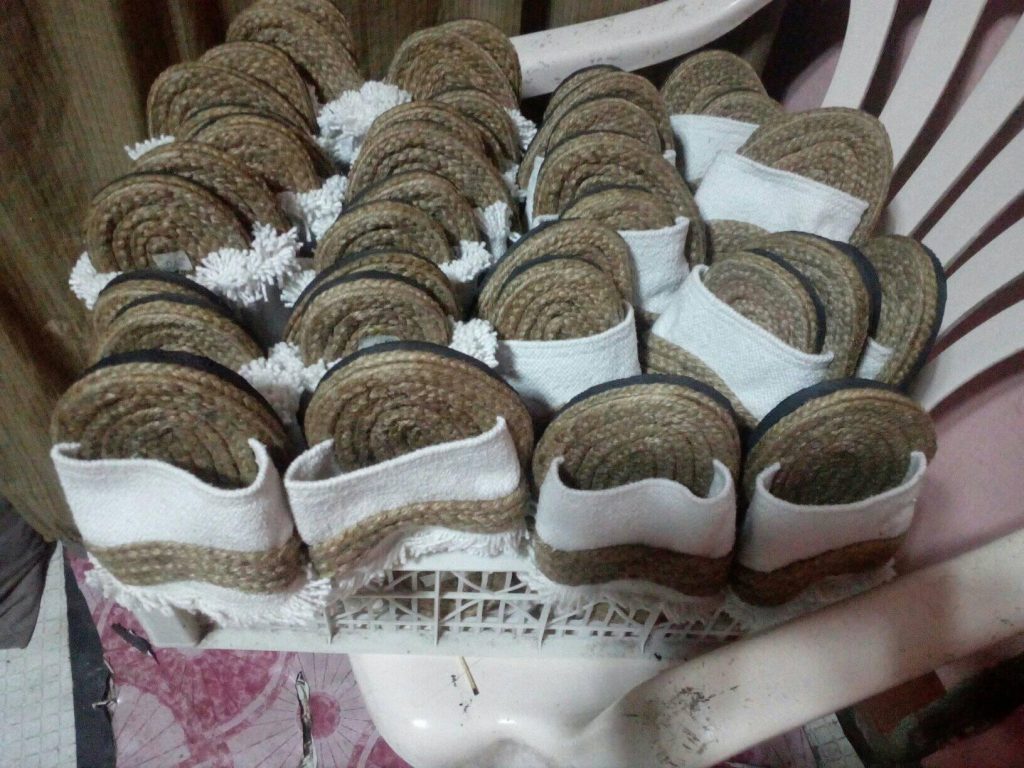 A collection of numerous handmade slippers is densely arranged in a white plastic container. The slippers have thick soles made from tightly coiled jute or a similar natural, coarse fiber, and they feature simple white fabric straps. Some of the slippers also have small, fluffy white pom-poms attached to the front. The close arrangement suggests the slippers are being stored or are ready for sale. Given the likely origin based on previous images and the current location of Taguig, Metro Manila, Philippines, these could be Filipino-made.