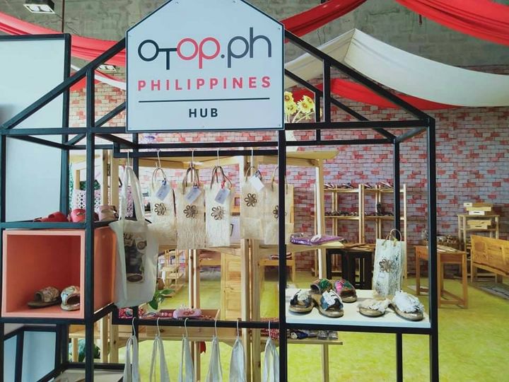 A retail display booth with a sign reading "otop.ph PHILIPPINES HUB" showcases various handcrafted items. The booth has a black metal frame resembling a stylized house. Hanging from the top are several light-colored tote bags with floral designs. Below, on shelves and tables made of light wood, are an assortment of slippers and other woven or textile products. The slippers feature different designs, some with floral patterns and others with simpler styles. In the background, a brick wall and draped red and white fabric decorations suggest a marketplace or trade fair setting. The "otop.ph PHILIPPINES HUB" sign indicates that the products are likely part of the One Town, One Product (OTOP) Philippines program, which promotes local crafts and goods from different regions of the Philippines. Therefore, the items displayed are likely Filipino-made.