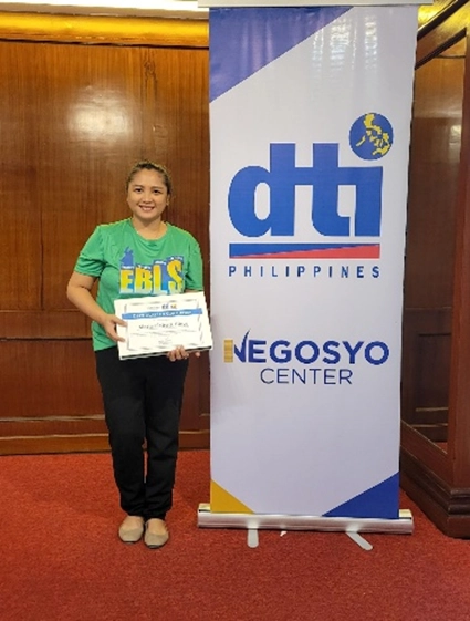 Marie Claire Bince of Purple Craft CDO smiles while holding her certificate from the DTI Enhanced Business Learning Session (EBLS), standing next to a DTI Philippines Negosyo Center banner.
