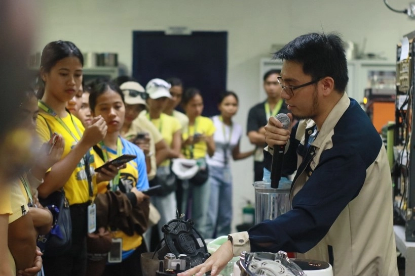 A DTI-BPS staff member holds a microphone and demonstrates testing procedures on small electrical appliances to a group of observing University of Northern Philippines (UNP) students inside the testing laboratory during their educational tour.