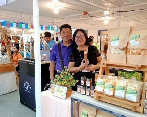 Noel and Myrna Hilario, the founders of Storehouse Food Products, are standing at a booth displaying their products, likely at a local trade fair or "pasalubong" center. Myrna is holding up one of their packaged goods. Various Storehouse products, including bags of chips and jars, are visible on the table and a small wooden shelf.