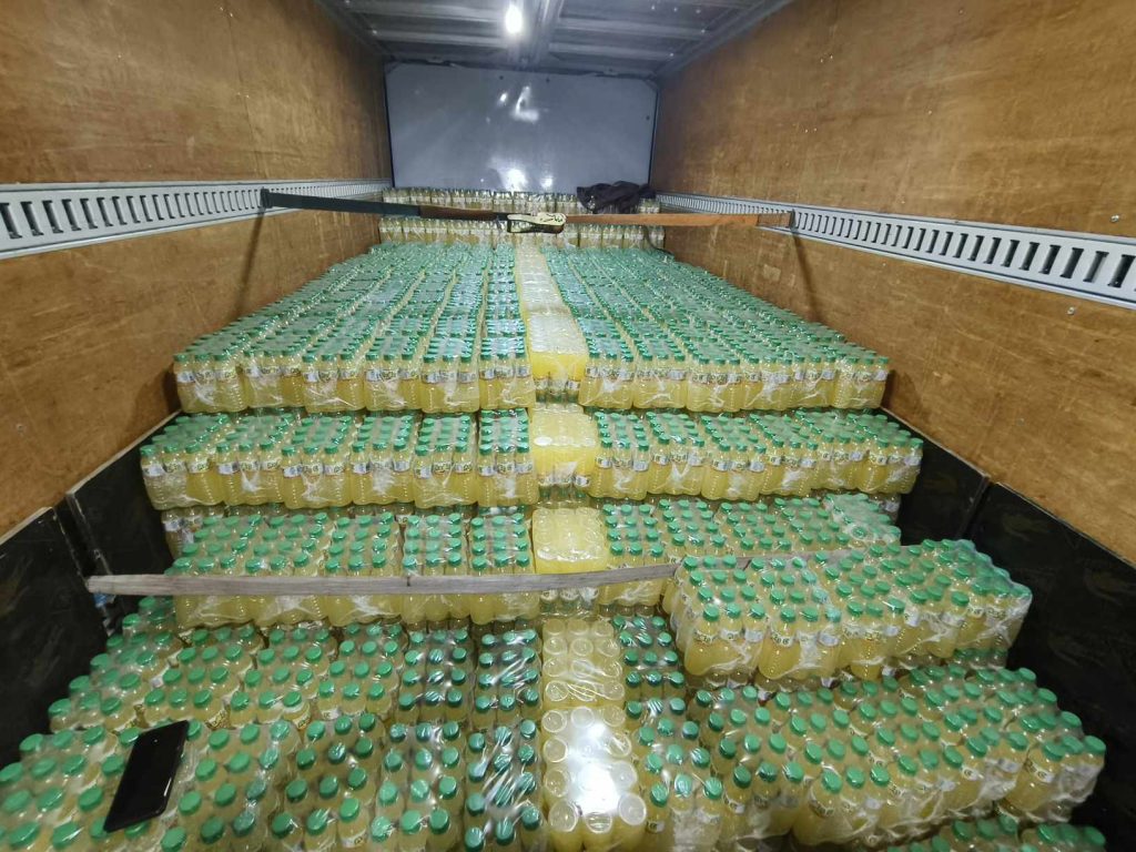 The interior of a truck is packed with numerous cases of yellow liquid bottles, possibly "AEON C" juice, stacked high and secured for delivery.