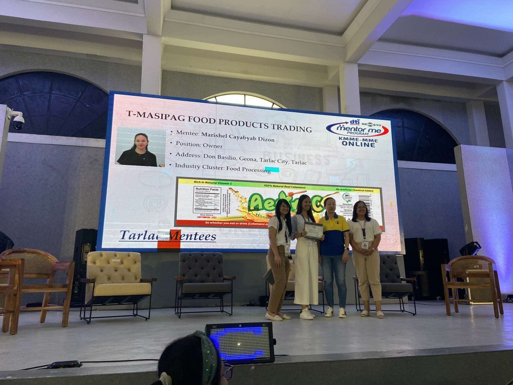 Four young women stand on a stage in front of a large screen displaying information about "T-MASIPAG FOOD PRODUCTS TRADING" and the "dti Negosyo Mentor Me Program KMME-MME ONLINE". The screen features a photo of a woman identified as the mentee, Marichel Cayabyab Dizon, along with details about her position, address in Tarlac City, Tarlac, and industry cluster (Food Processing). The women on stage appear to be holding certificates or awards. The setting suggests a graduation or recognition ceremony for participants of the mentorship program. Given the location context of Tarlac and the program's affiliation with the Philippine Department of Trade and Industry (DTI Negosyo), the individuals on stage are likely Filipina.