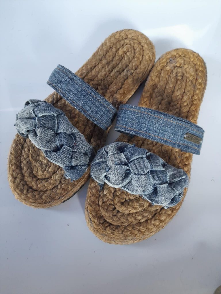 A pair of handmade slippers is presented on a light gray surface. The slippers feature thick soles made from tightly coiled jute or a similar natural, coarse fiber. Each slipper has two straps made of blue denim fabric. One strap is a simple band, while the other is wider and appears to be woven or braided, creating a textured pattern. The slippers have a rustic and artisanal look. Given the likely origin based on previous images and the current location of Taguig, Metro Manila, Philippines, these could be Filipino-made.