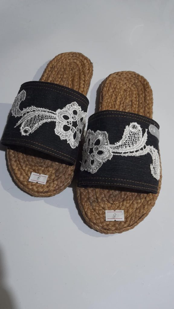 A pair of handmade slippers is displayed on a light gray surface. The slippers have thick, textured soles made from tightly coiled jute or a similar natural fiber. The straps are made of a dark-colored fabric, possibly black denim, and are decorated with intricate white floral embroidery or lace-like patterns. Small white tags with numbers are attached to the soles of each slipper. The slippers appear to be artisanal and have a unique, decorative style. Given the likely origin based on previous images and the current location of Taguig, Metro Manila, Philippines, these could be Filipino-made.