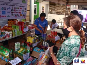 An outdoor scene showing several people, who appear to be of Southeast Asian descent based on visual cues, at a market or promotional event. A woman wearing a green patterned top and a black face mask is in the foreground, holding a plastic bag and looking at items on a table. The table is filled with various packaged goods, including food items and beverages. A man in a blue shirt is standing behind the table, possibly organizing or assisting with the display. A young boy is seated at the table, looking down. A sign in the background indicates hours of operation and contact numbers, suggesting this is a sales event. A logo for 'dti' is visible in the lower right corner, indicating a connection to the Philippine Department of Trade and Industry. The overall setting appears to be a busy, outdoor market or promotional event, potentially the 'Diskwento Caravan' mentioned in the caption. The presence of numerous packaged goods suggests a focus on retail or consumer products.