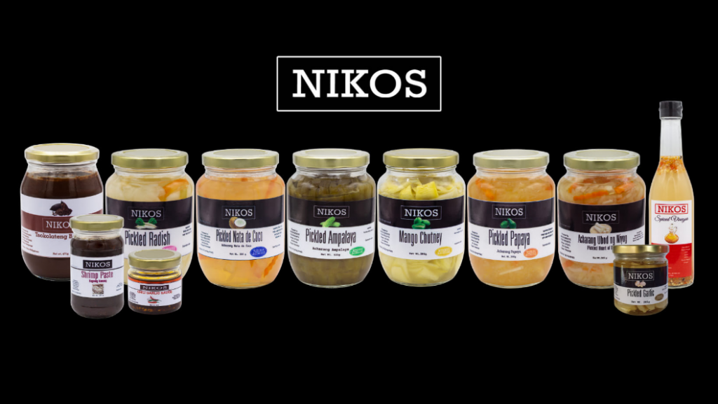 A product shot displays a collection of "NIKOS" brand pickled food items against a dark background. The central focus is on eight glass jars and one glass bottle, each containing different pickled contents and labeled with the "NIKOS" logo and the specific product name. From left to right, the visible labels include: "Nikos Tsukemono Pickled Radish", "Pickled Nata de Coco", "Pickled Ampalaya", "Mango Chutney", "Pickled Papaya", "Atcharang Thai Chili Relish", and "Pickled Garlic". The bottle on the far right also bears the "NIKOS" label. The variety of colors and textures within the jars suggests a range of pickled fruits and vegetables. The overall presentation is clean and emphasizes the "NIKOS" brand and its diverse line of pickled products. Given the likely origin of the brand based on previous images, these products are likely from the Philippines.