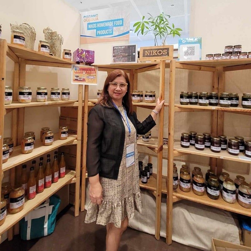 A woman stands smiling in front of a wooden display shelf filled with various jars and bottles of food products. A banner above reads "NIKOS HOMEMADE FOOD PRODUCTS". The woman is wearing glasses, a patterned dress, and a dark sheer jacket, and she has a name tag hanging around her neck. The shelves are stocked with items that appear to be preserved foods, sauces, or condiments. A small potted plant sits on the top shelf. The setting suggests a trade show or a small business showcasing its products. Based on the branding, the business is likely named "Nikos" and specializes in homemade food items.